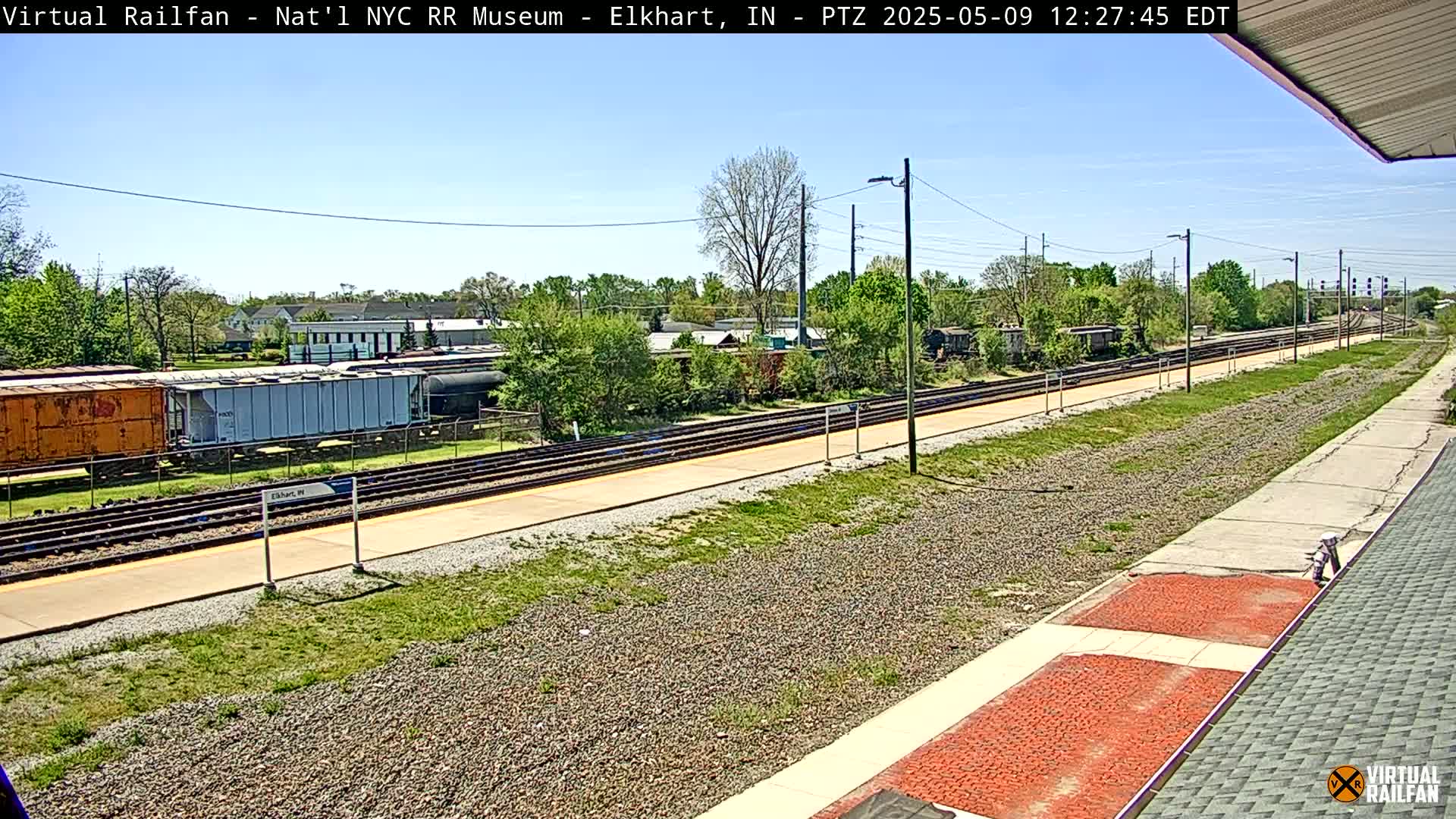 Under a clear blue sky, railroad tracks stretch into the distance past several freight cars and a station platform, bordered by green grass and trees.