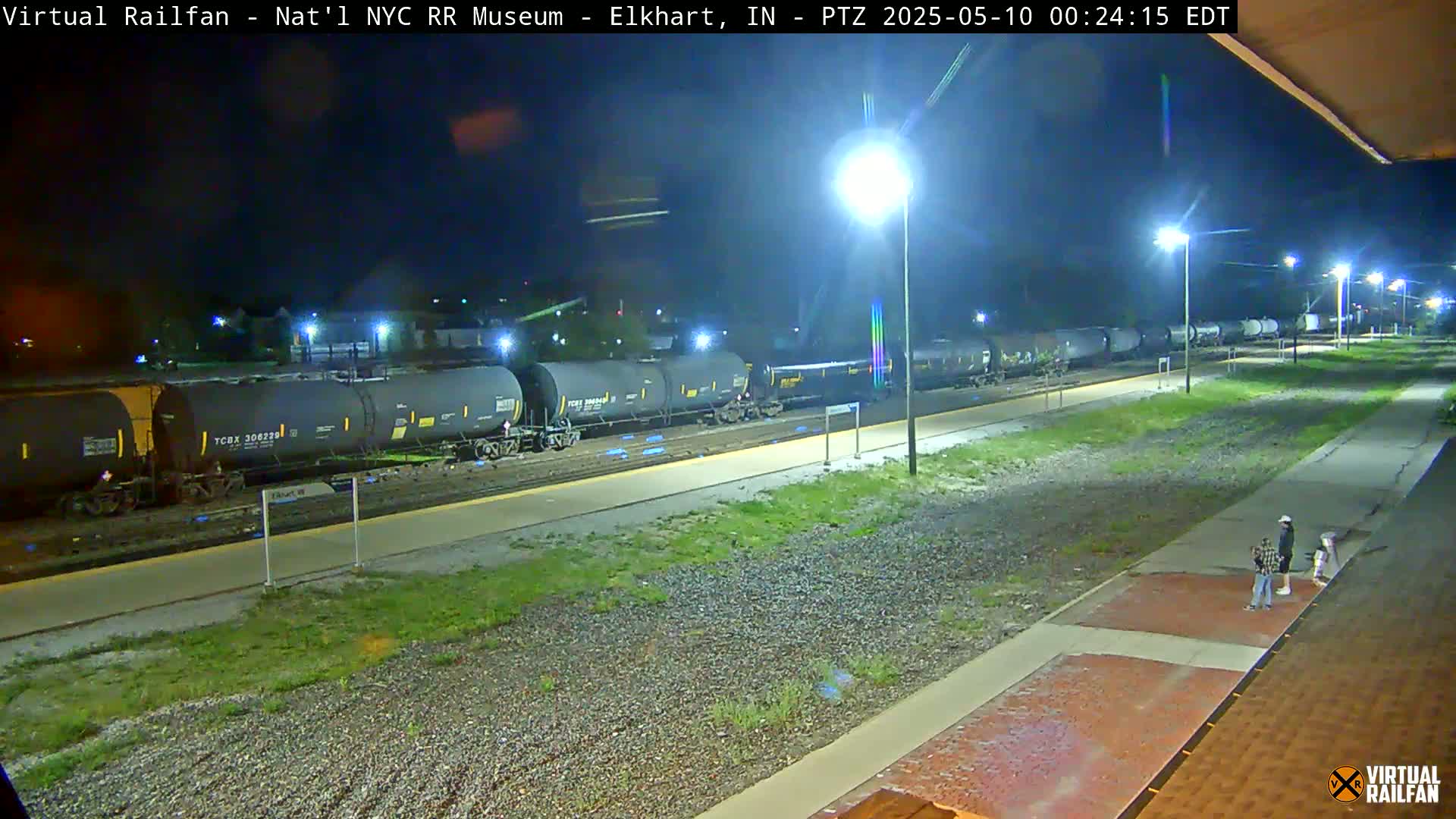 A long freight train of dark tanker cars sits at a station at night under bright lights, with a few people standing on a nearby sidewalk.