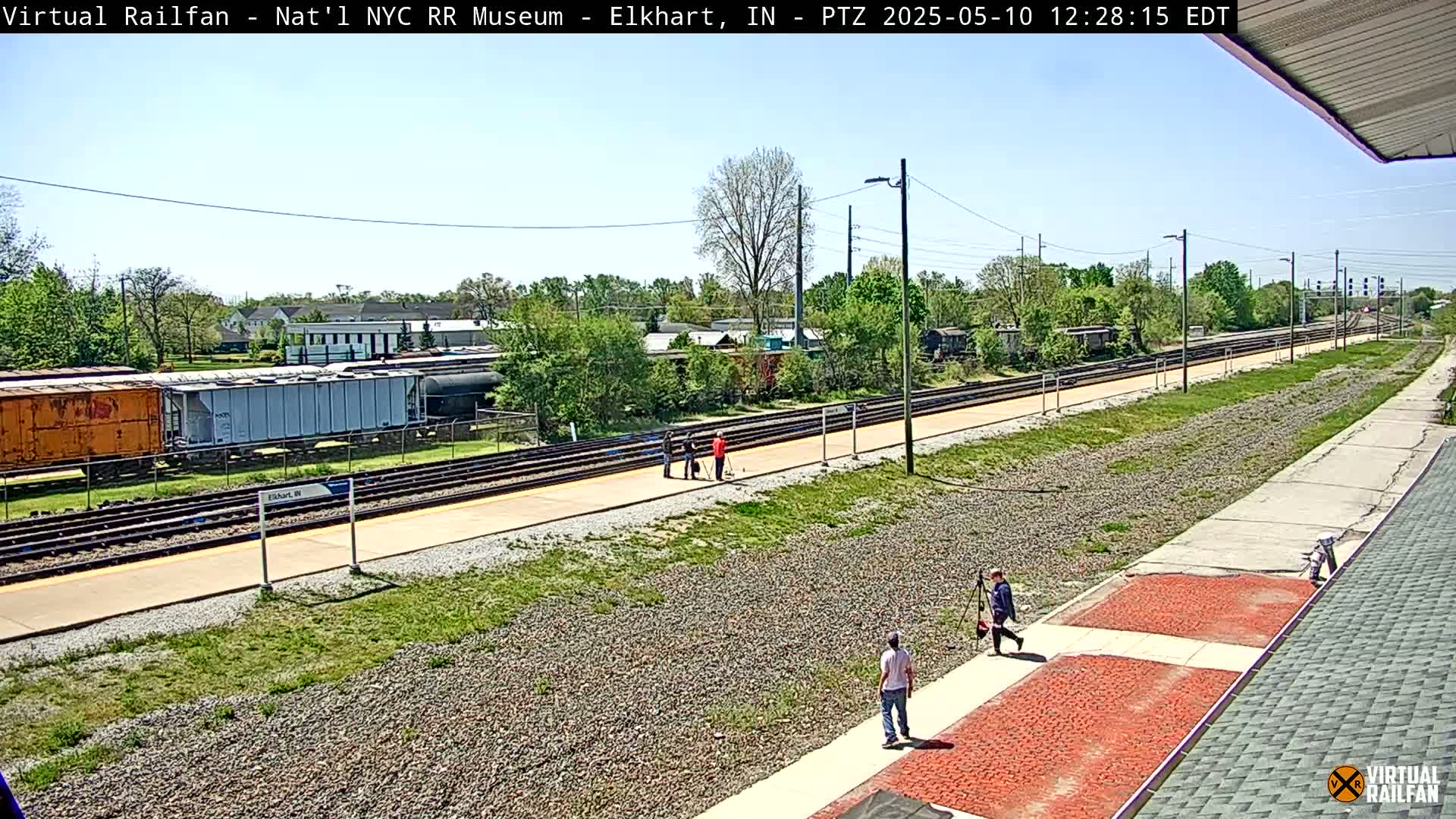 A sunny day at a train station shows several people walking along a paved path next to train tracks with boxcars and other rail cars visible in the distance.
