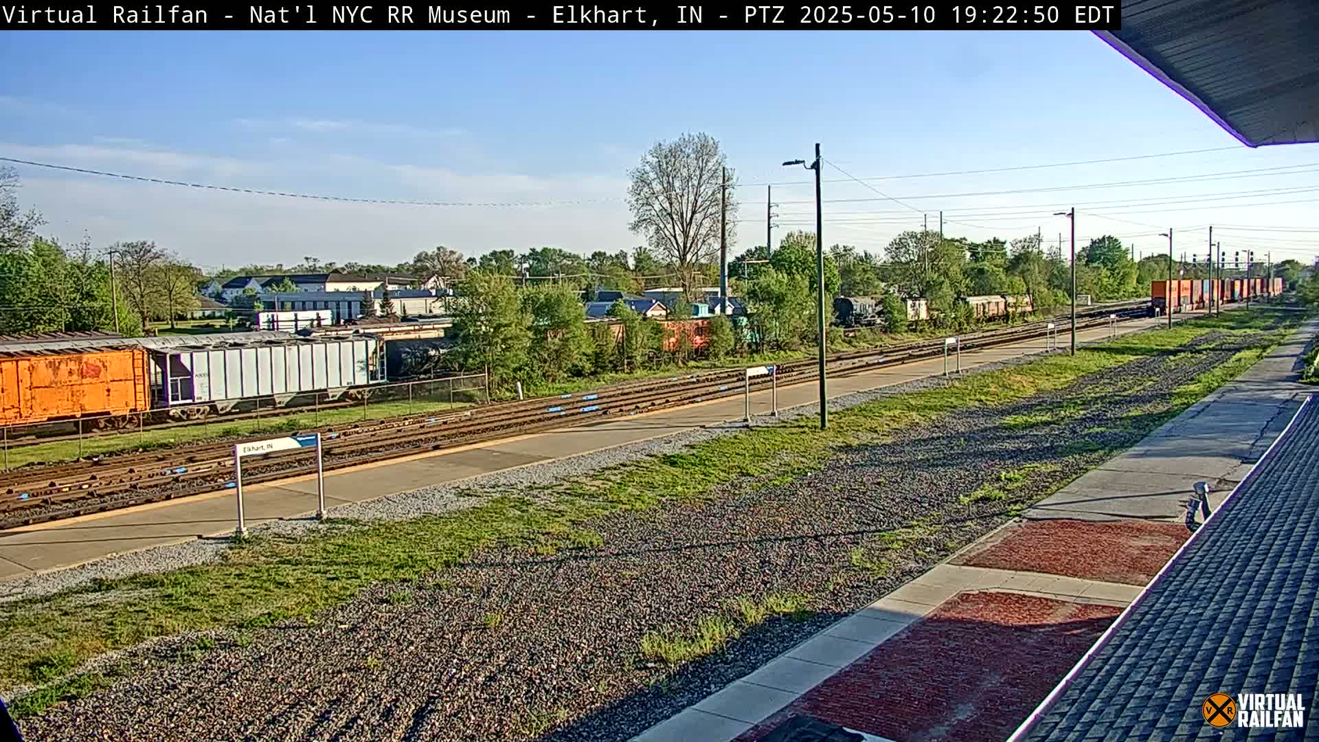 A mostly sunny daytime view of railway tracks with several freight cars and a platform, surrounded by green vegetation.