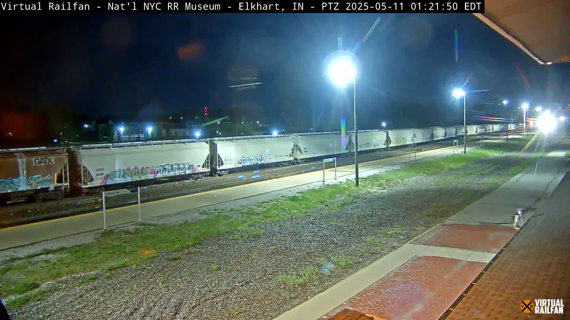 A long freight train with graffiti on some of the cars sits at night on a railway platform under bright lights.
