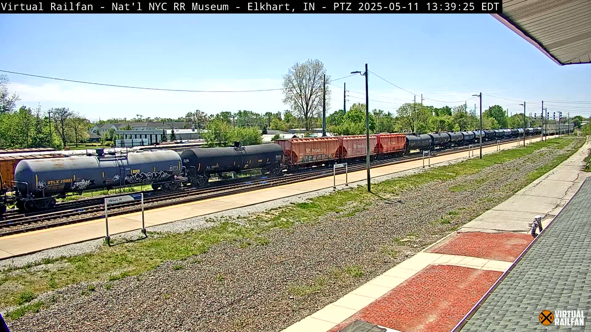 A long freight train of tanker cars and boxcars sits on a track next to a platform on a sunny day.