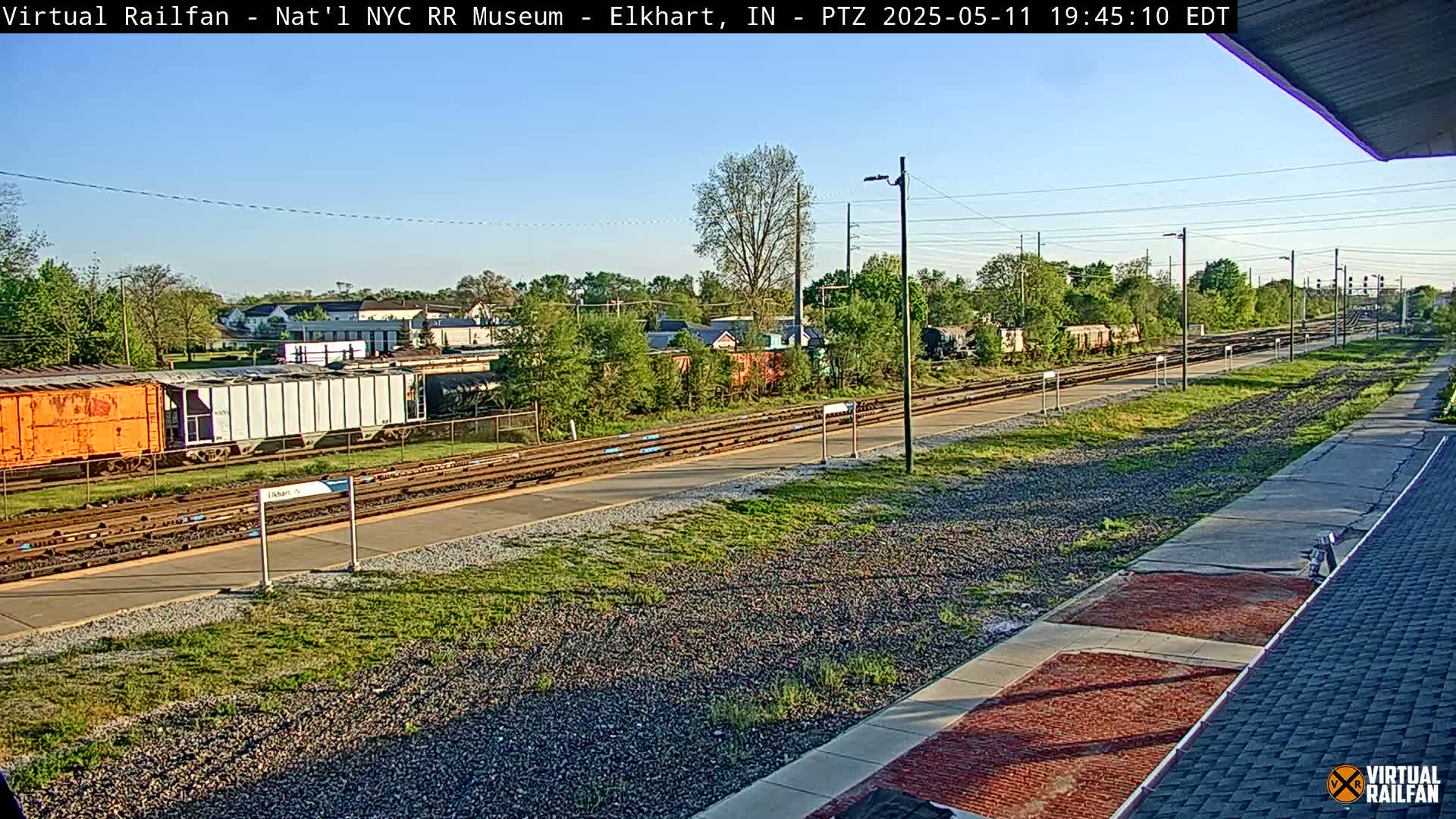 A sunny view of a train station with several railway tracks, freight cars, and overgrown vegetation.
