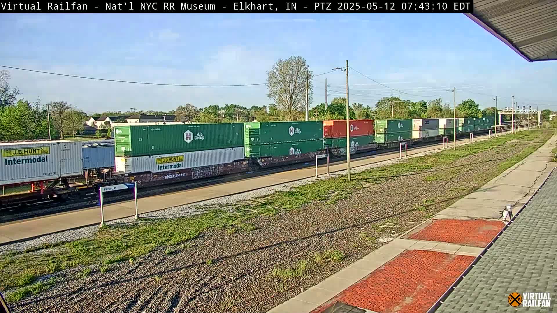 A long freight train carrying many green and orange shipping containers sits on a railway track under a partly sunny sky.