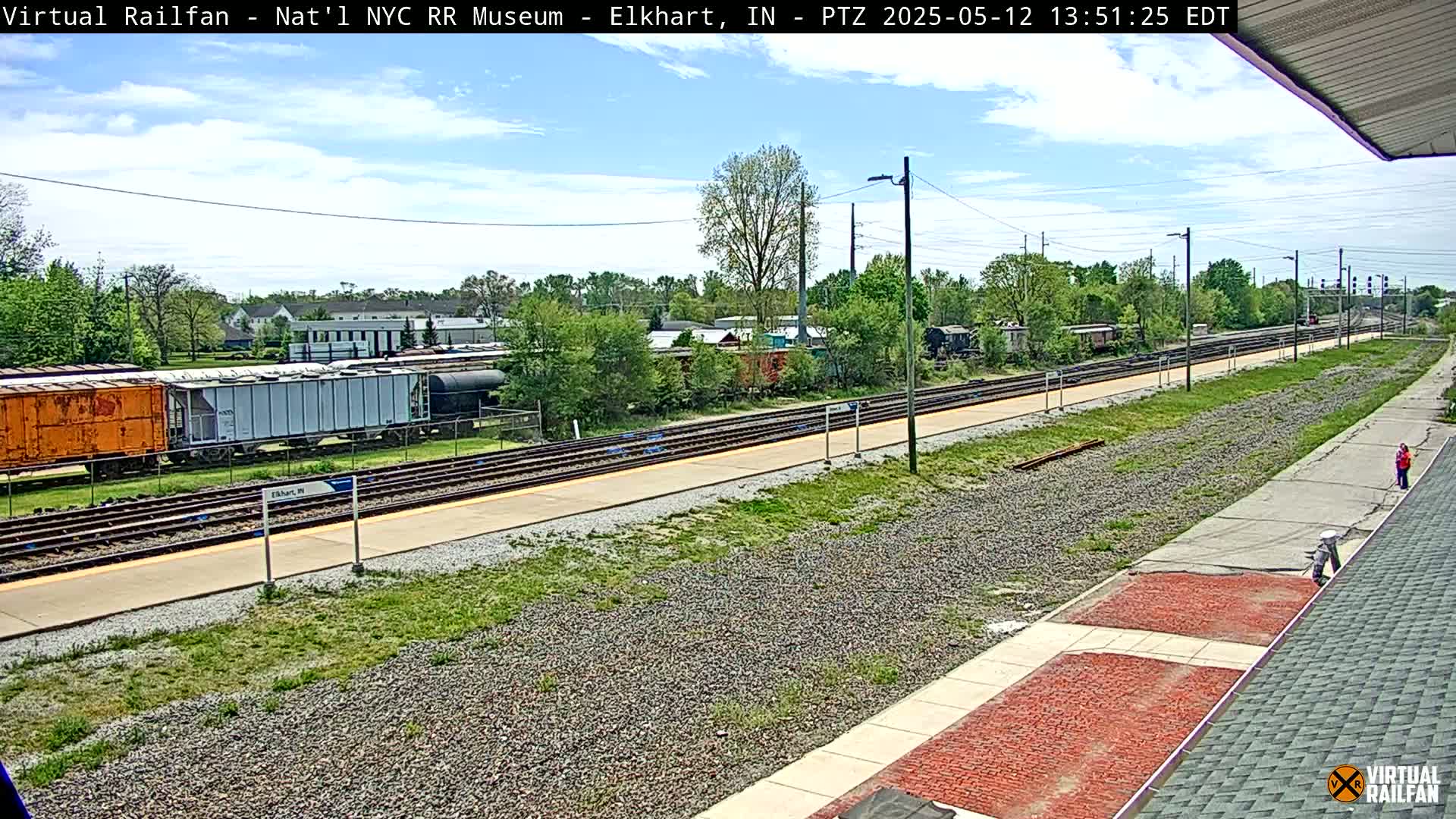 A mostly sunny day at a train station shows several freight cars and a platform with a single person standing nearby.