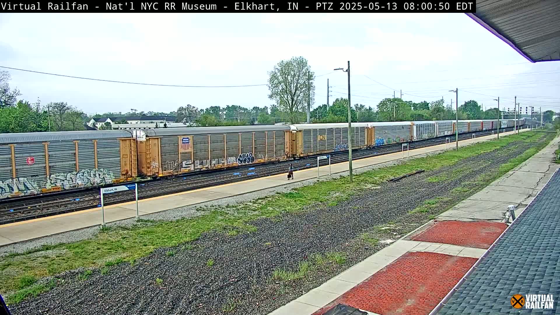 A long freight train with graffiti on some of its cars sits at a station on a partly cloudy day.