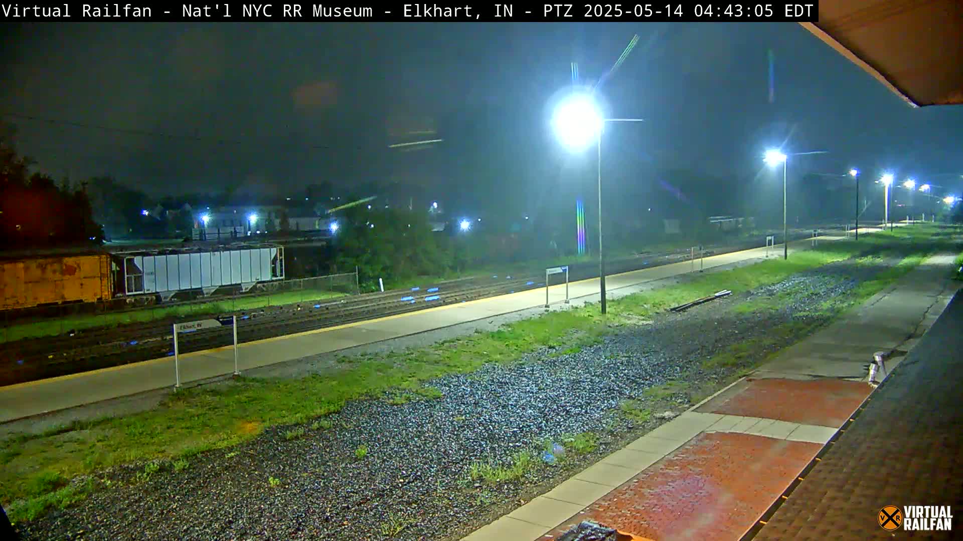 A nighttime view of a rain-slicked train platform, with train cars and tracks visible in the background under the glow of several streetlights.