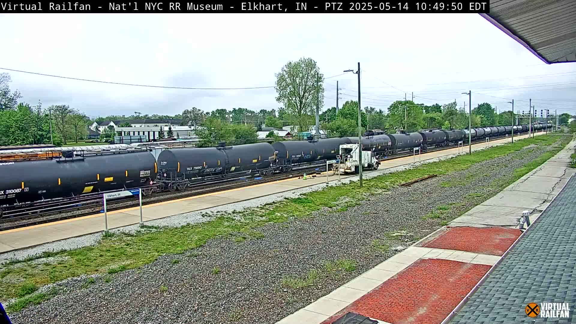 A long train of black tanker cars sits at a station on a partly cloudy day.