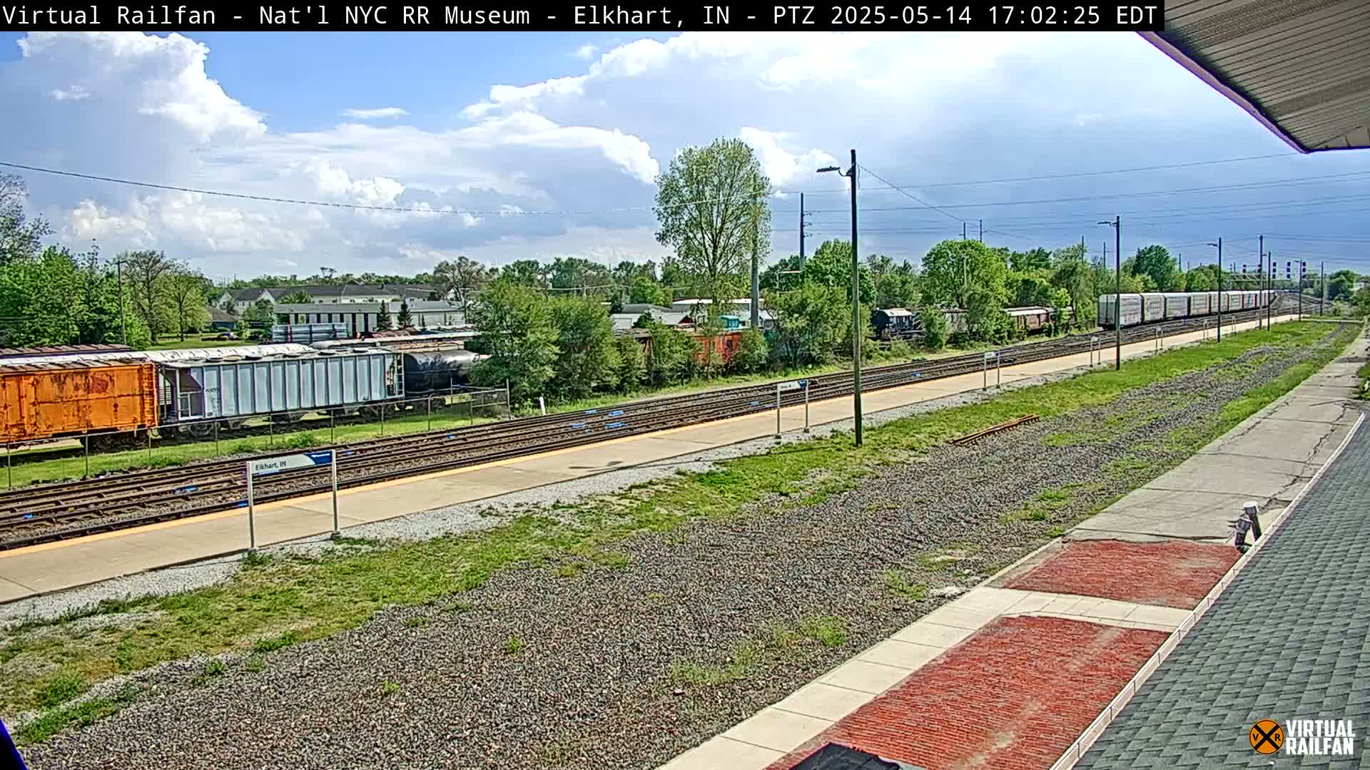 A partly cloudy sky overlooks a train station with several freight cars and a long train on the tracks.