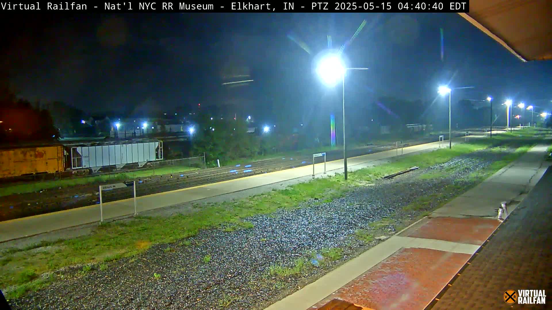 A nighttime view of a train platform with a few freight cars visible in the distance, illuminated by bright lights.