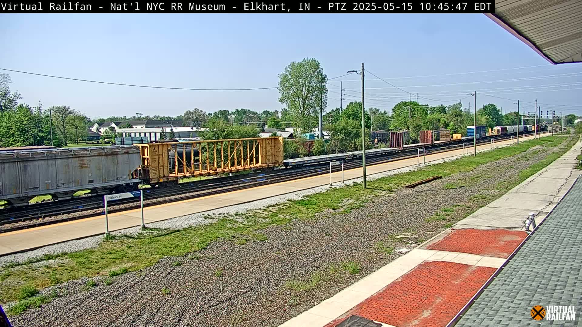 A long freight train is visible on a sunny day, passing by a train station platform with grassy areas on either side of the tracks.