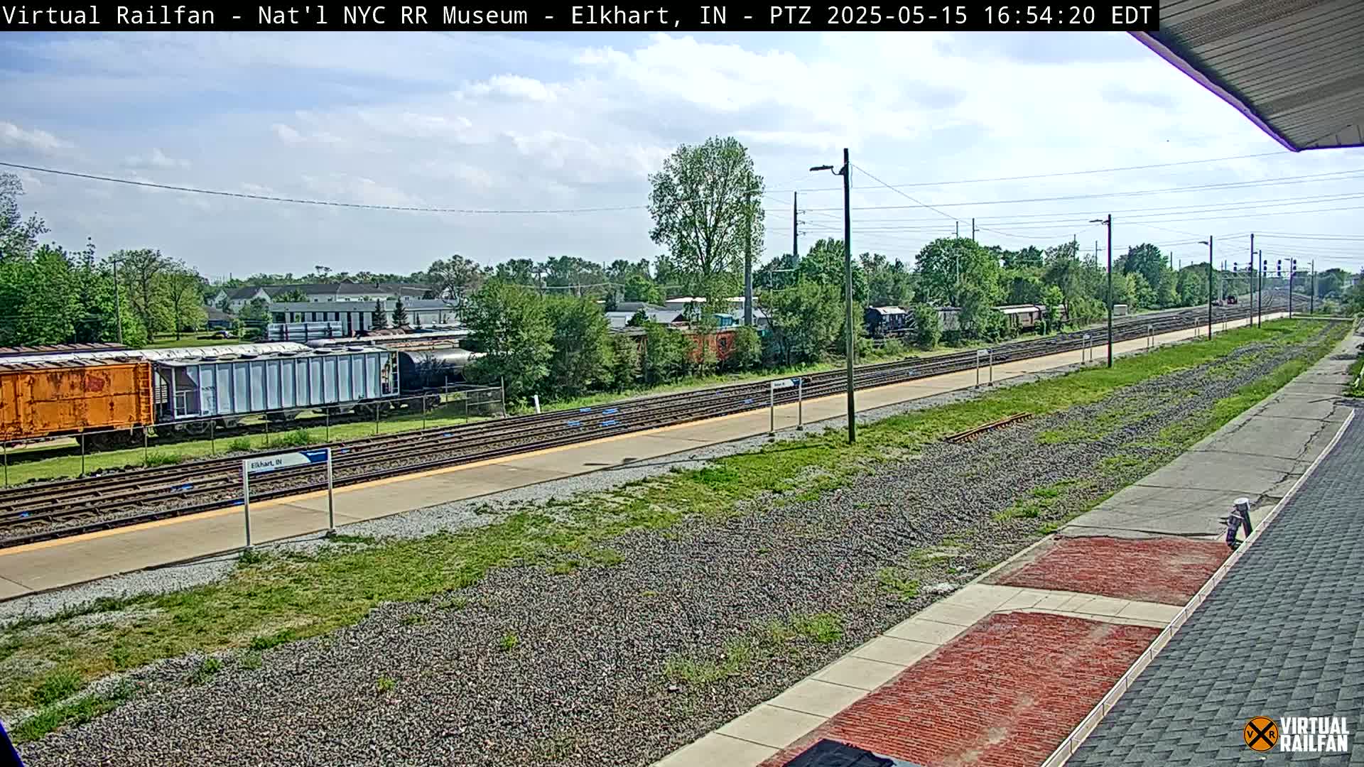 A mostly sunny day reveals a railway scene with several freight cars parked near a platform and multiple train tracks extending into the distance, surrounded by green vegetation.