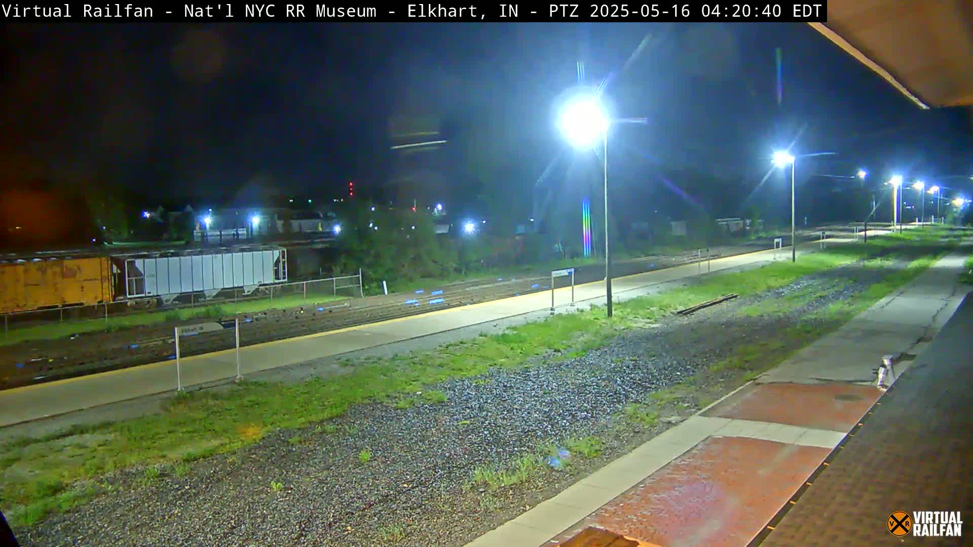 A nighttime view of a train station platform with railway tracks, some freight cars, and several bright lights illuminating the scene.