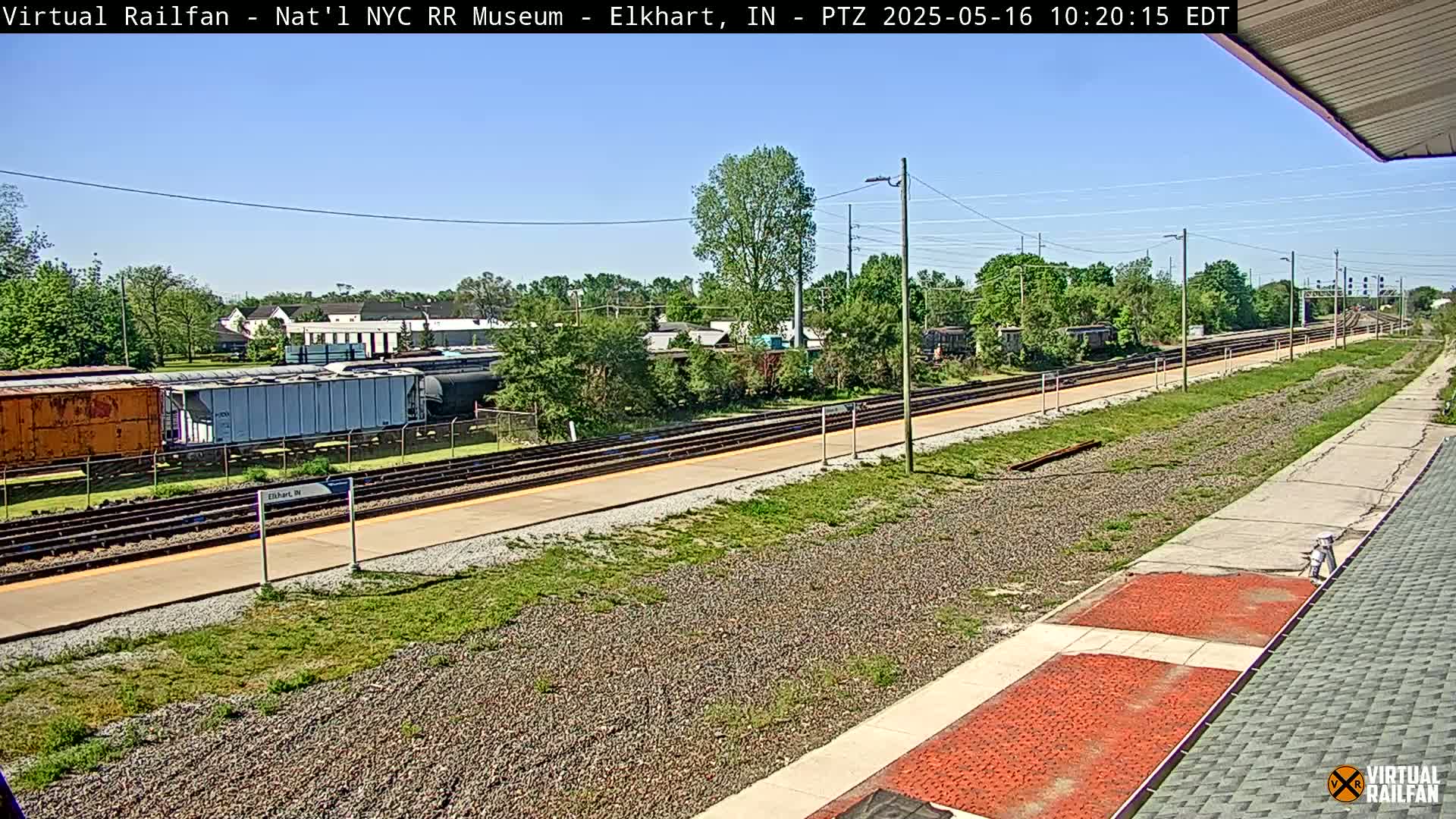 A sunny day reveals a train platform with railway tracks extending into the distance, alongside parked rail cars and lush green vegetation.