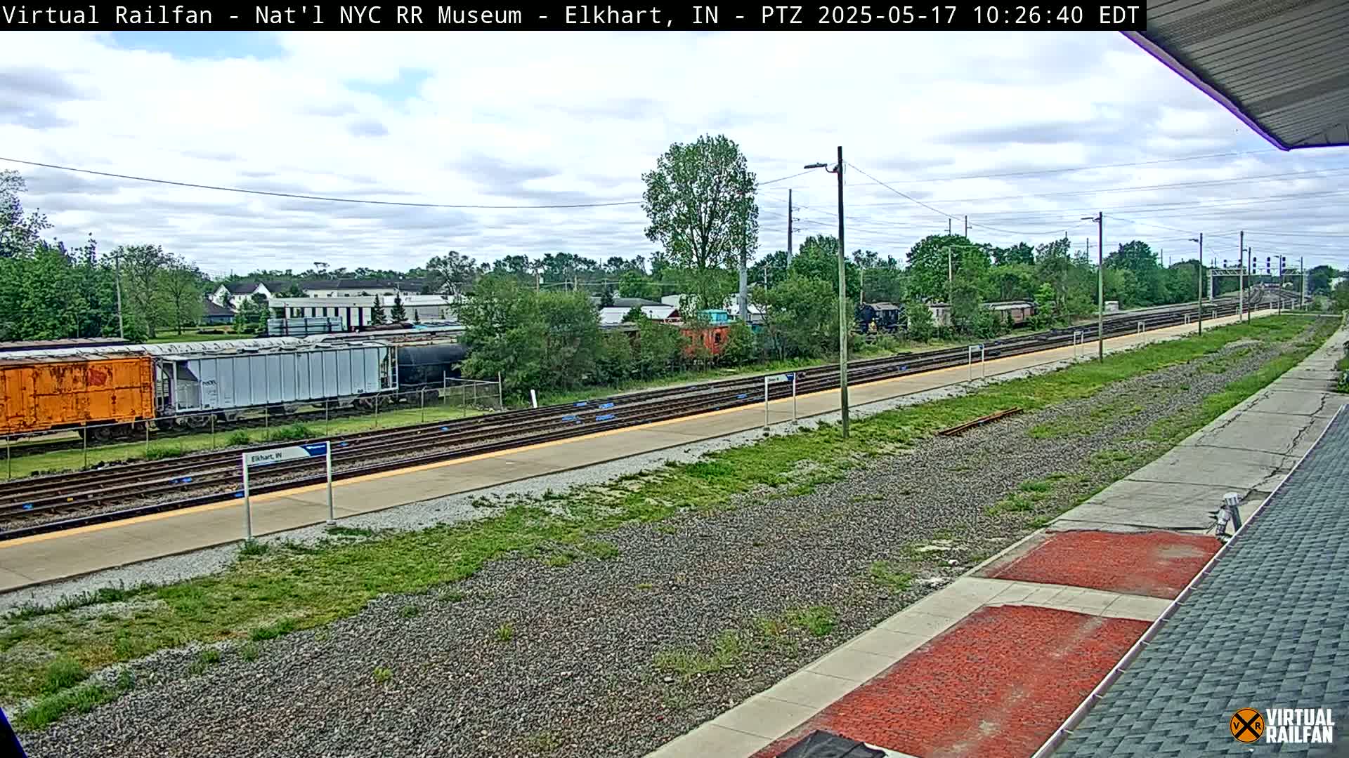A train station platform on a partly cloudy day, with multiple train cars visible on the tracks and lush green vegetation surrounding the area.
