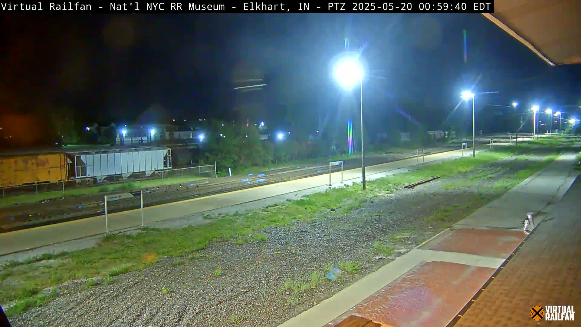 A nighttime view of a train platform and tracks, illuminated by streetlights, with several railway cars visible in the background.