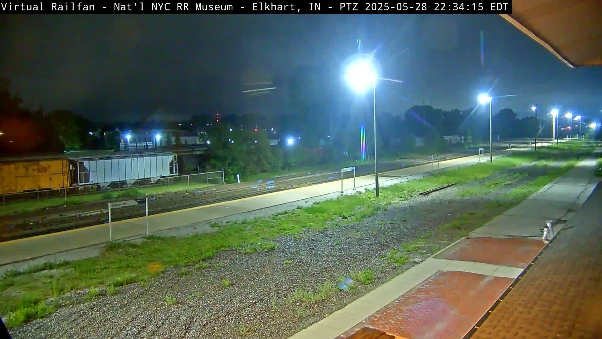 A nighttime view of a train station platform with railway tracks, some freight cars, and several lights illuminating the scene.