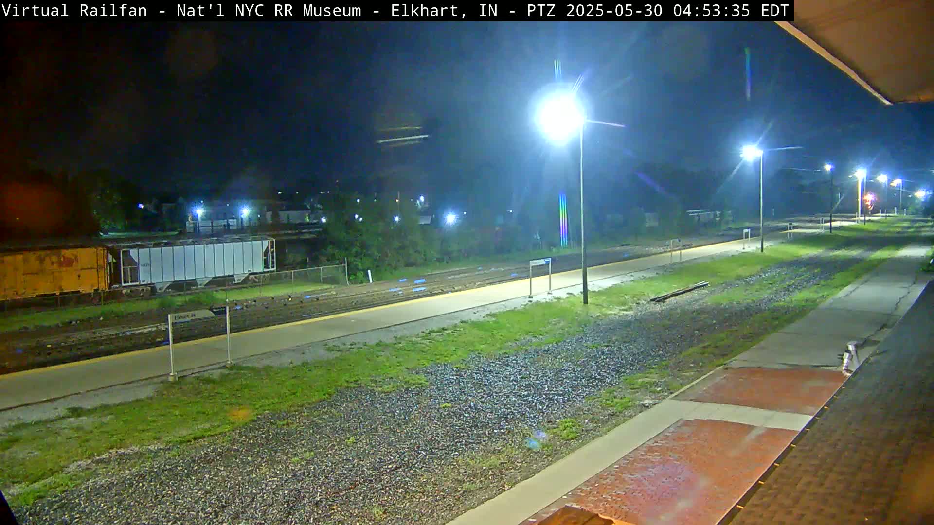 A nighttime view of a train platform and railway tracks, with several freight cars visible in the distance under a dark sky illuminated by streetlights.
