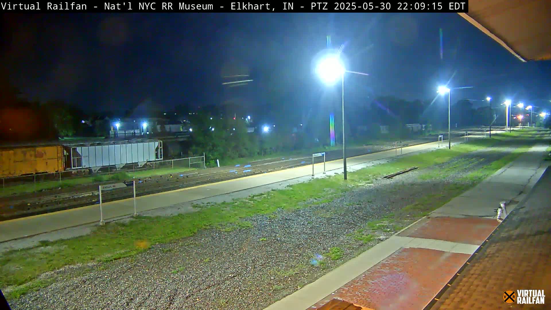 A nighttime view of a train station platform and rail tracks, illuminated by several streetlights.