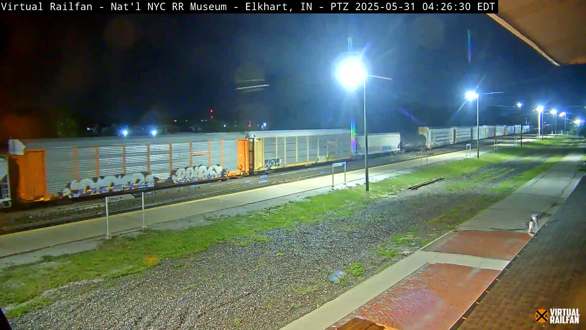 A long freight train, some cars of which are graffitied, sits at a station at night under several bright lights.