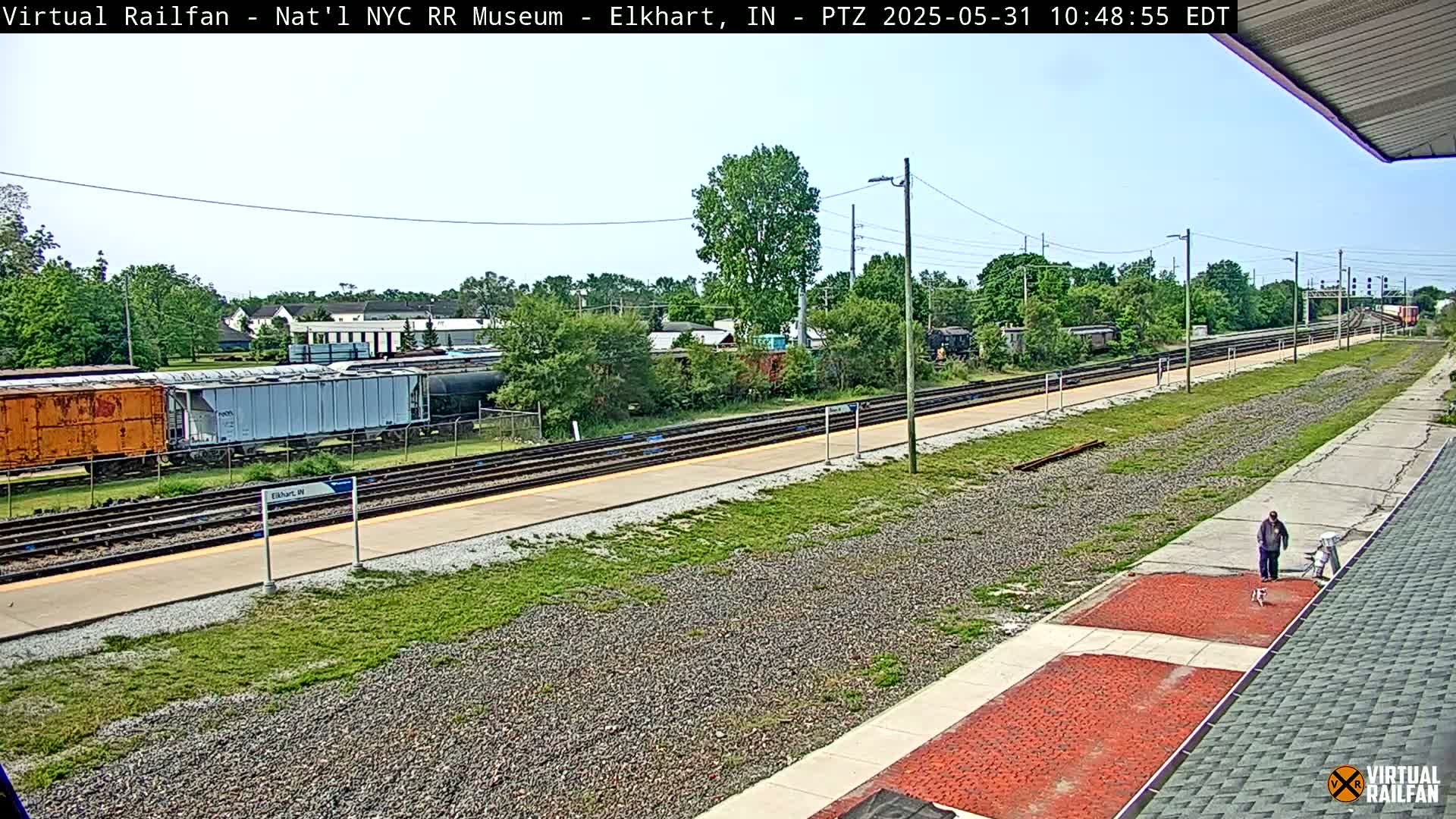 A sunny day at a train station shows several railcars, a person walking a dog, and multiple train tracks extending into the distance.