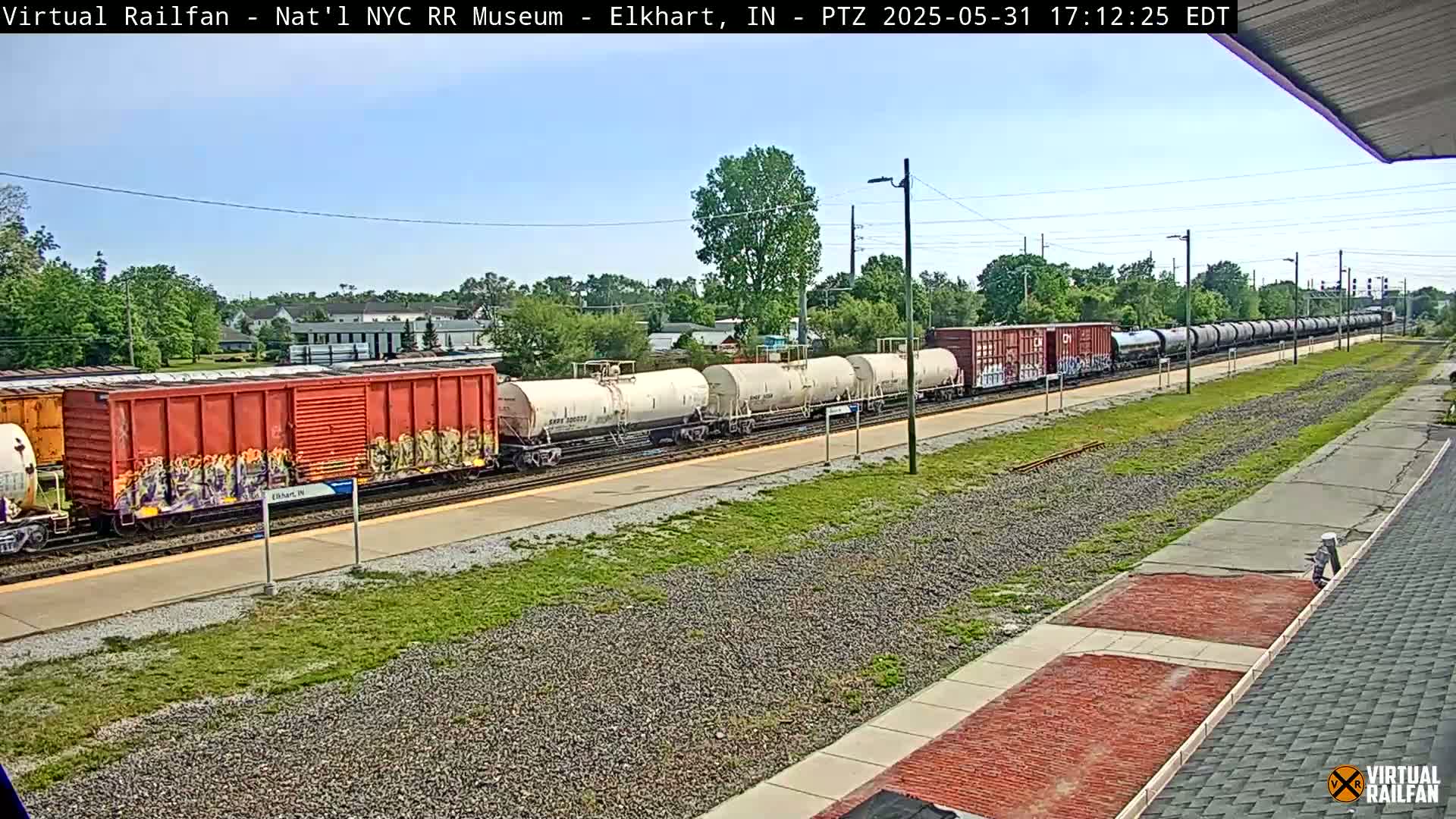 A long freight train, consisting of boxcars and tanker cars, some of which are graffitied, is seen on a sunny day.