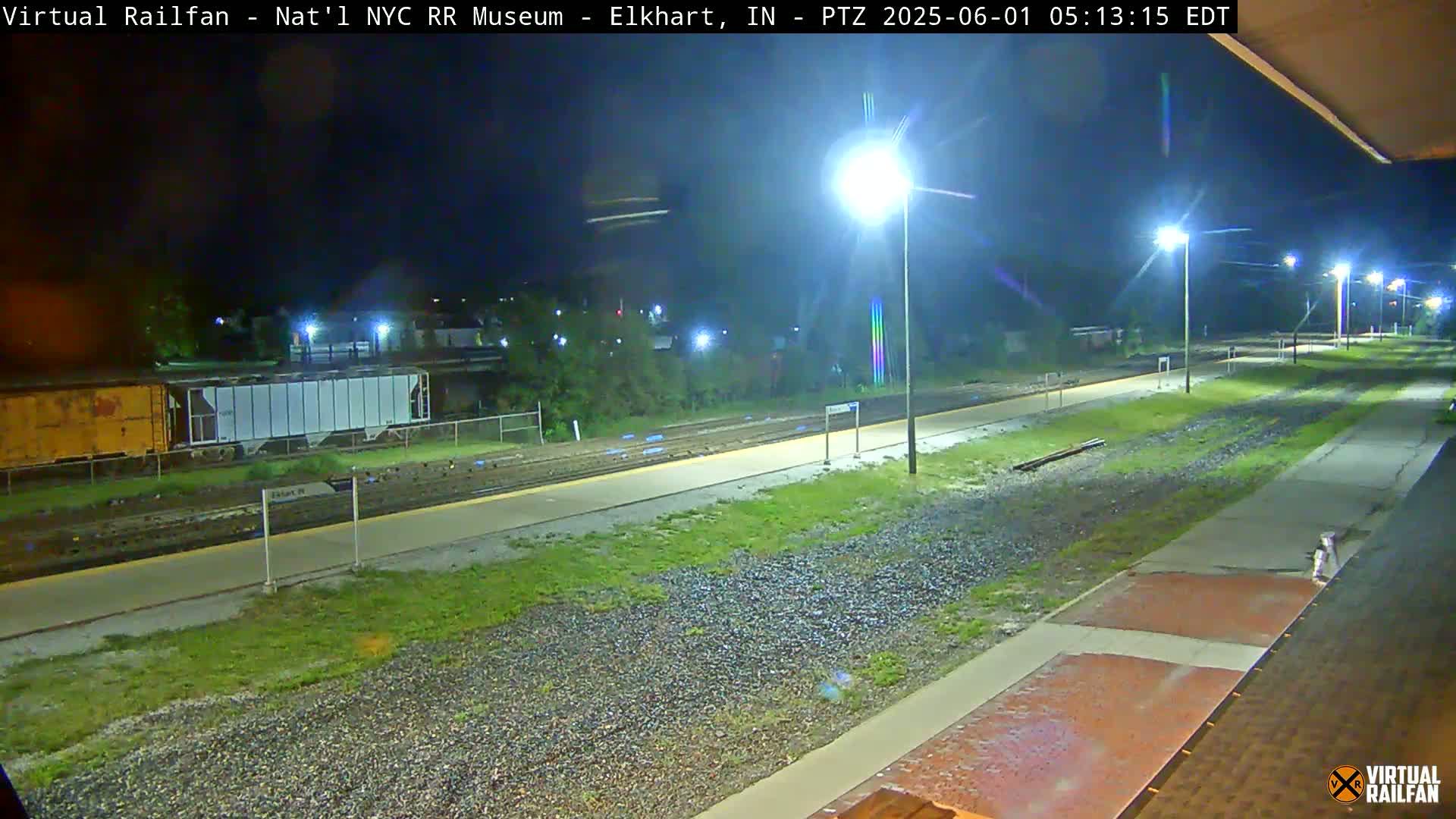 A nighttime view of a train platform and railway tracks, illuminated by several streetlights, with a train car visible in the background.
