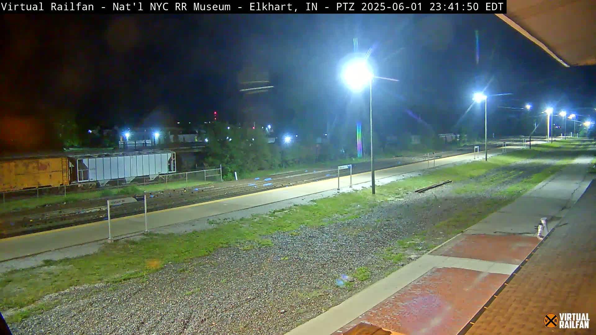 A nighttime view of a train station platform and railway tracks, illuminated by several bright lights.