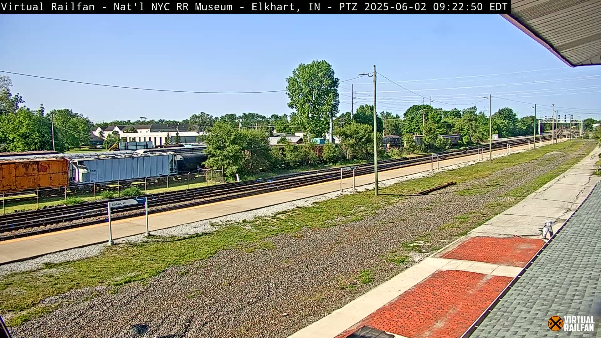 A sunny day at a train station shows several freight cars parked on a track, with a platform and grassy areas nearby.