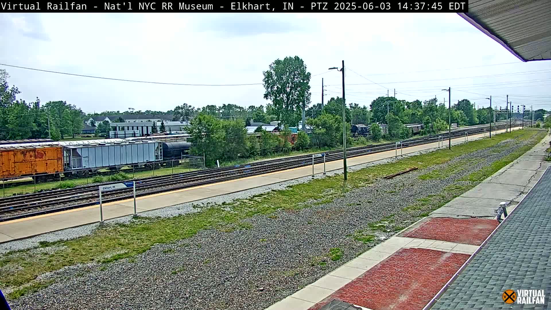 A mostly sunny day at a train station shows several freight cars and railway tracks, with a grassy area and buildings in the background.