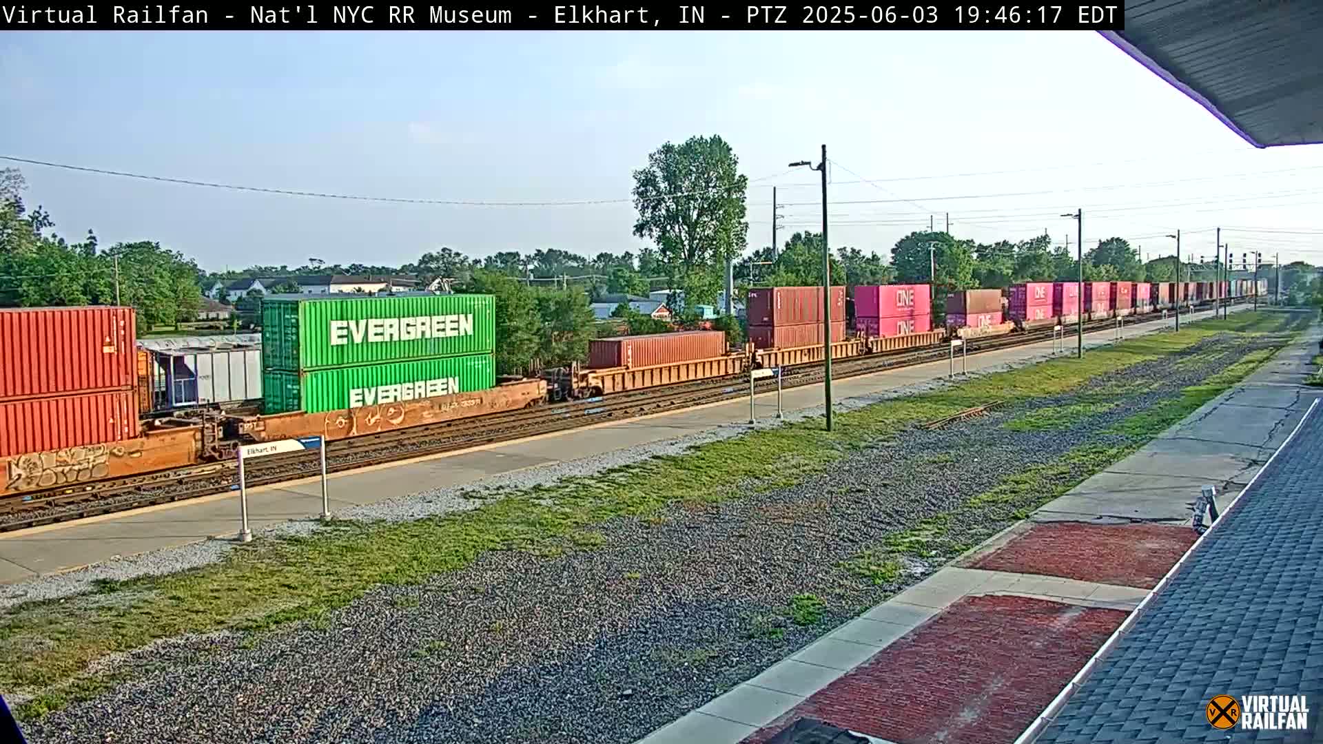 A long freight train carrying various colored shipping containers sits on a railway track under a partly sunny sky.