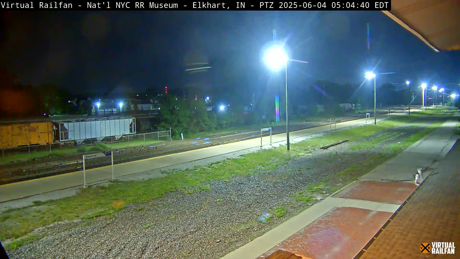 A nighttime view of a train platform and tracks, illuminated by several streetlights, with rail cars visible in the distance.