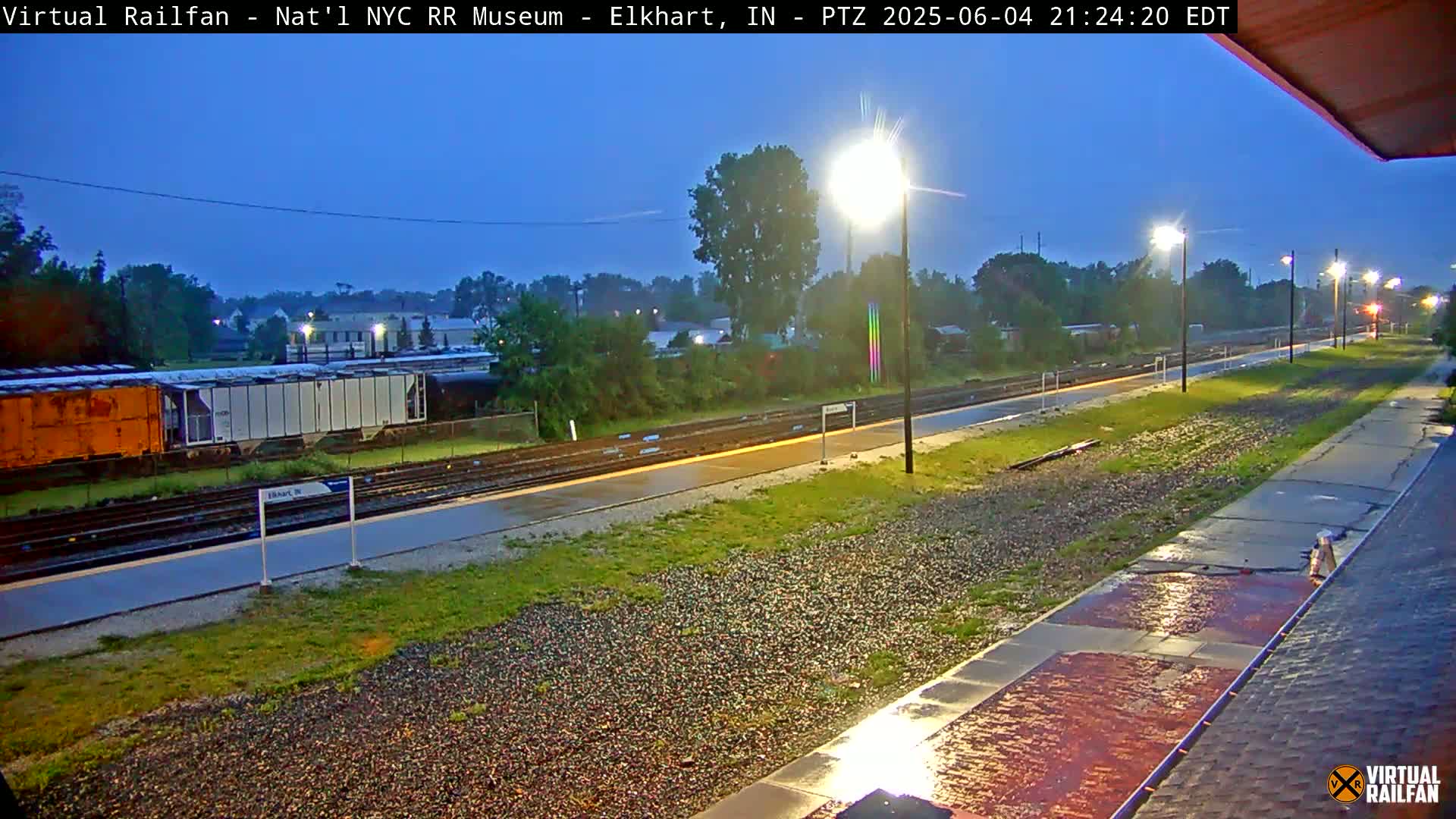 A rain-slicked train platform at night, with several railcars visible in the background under a twilight sky.