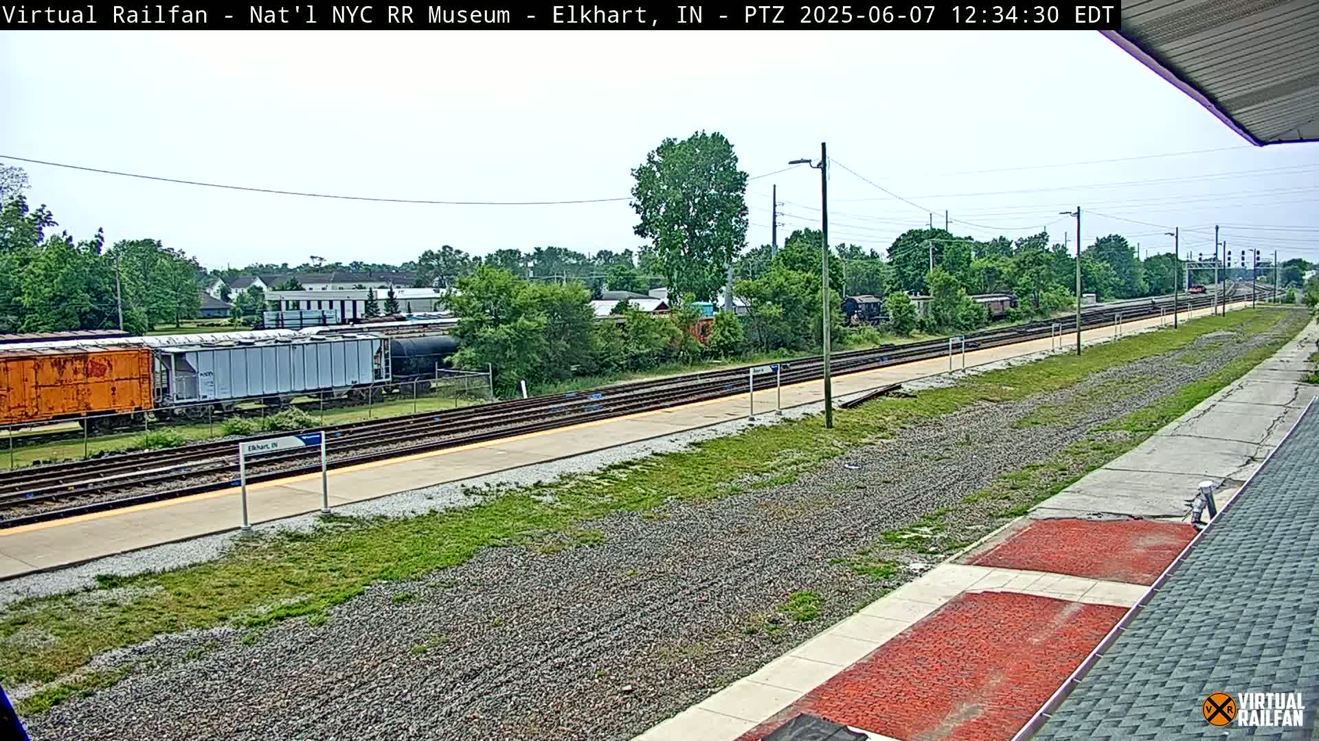 A sunny day at a train station shows several rail cars and tracks, with a grassy area and some buildings in the background.