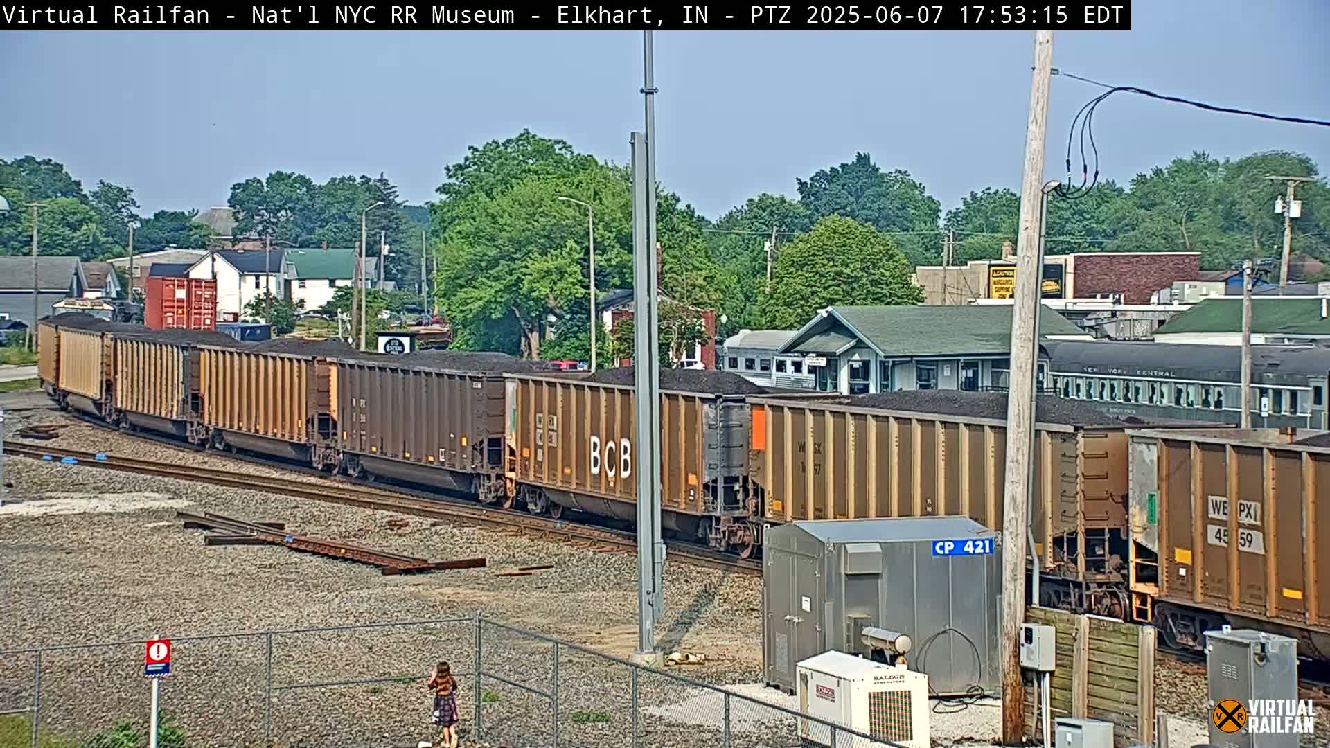 A long freight train carrying coal is passing through a small town on a sunny day.