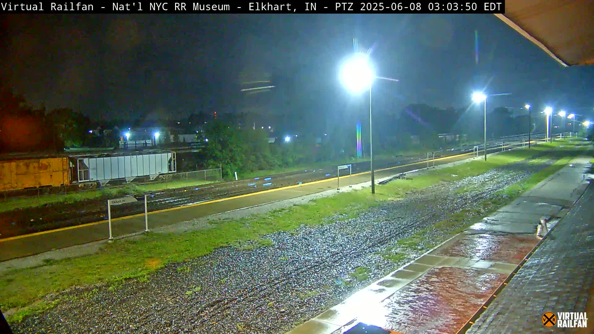 A nighttime view of a rain-slicked train platform and tracks, illuminated by several streetlights.