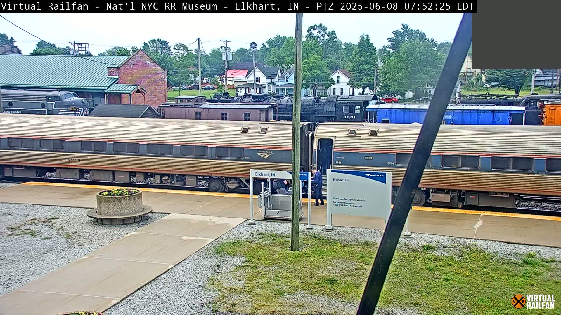 A long, gold Amtrak passenger car sits at a train station on a partly cloudy day, with several other train cars and locomotives visible in the background.