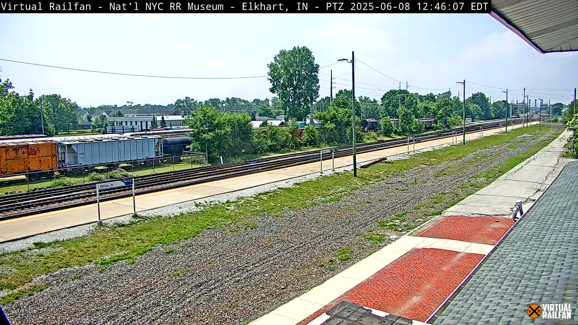 A sunny day at a train station with railway tracks, several railcars, and grassy areas surrounding the platform.