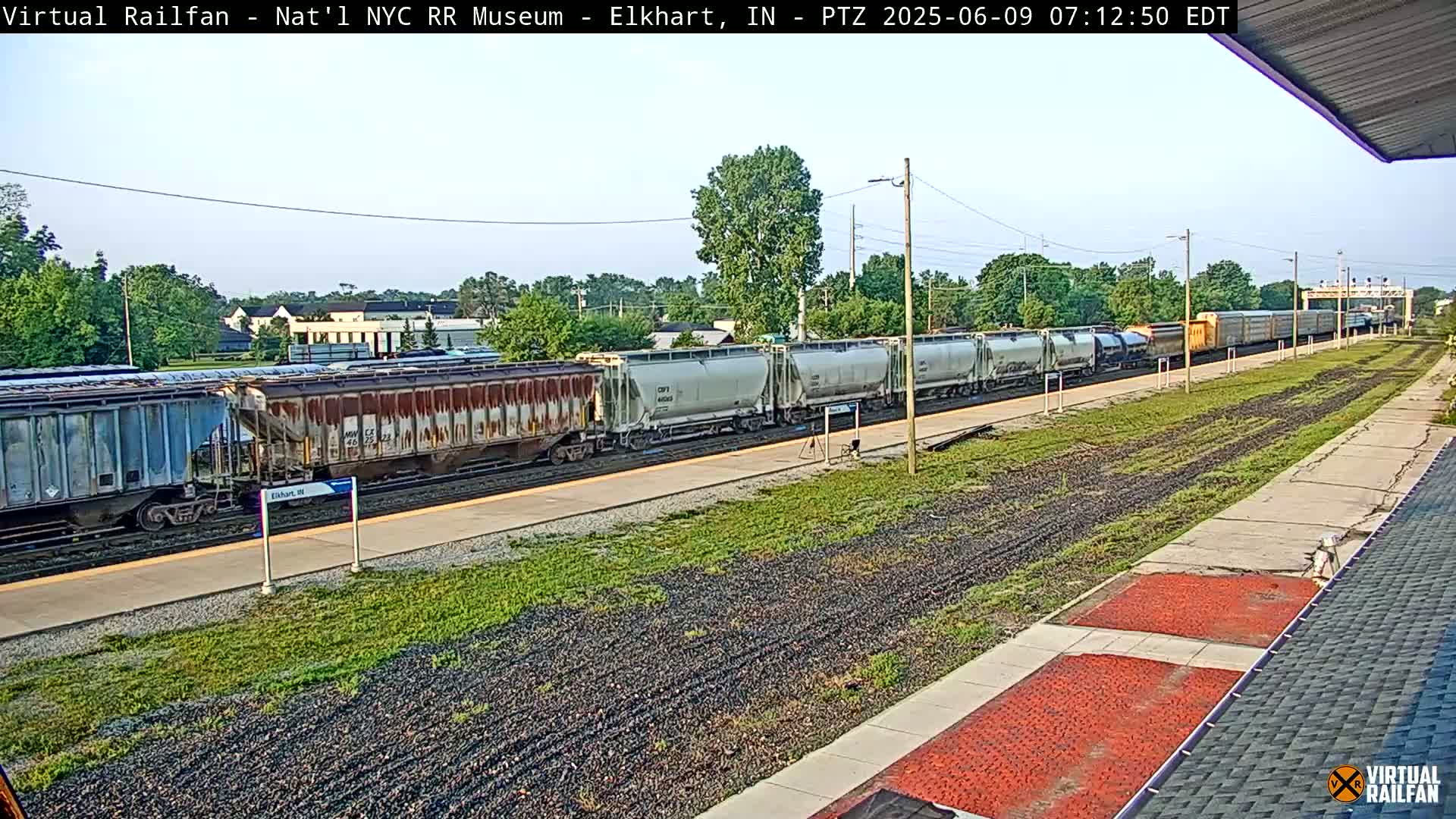 A long freight train of various colored cars passes through a train station on a partly sunny day.