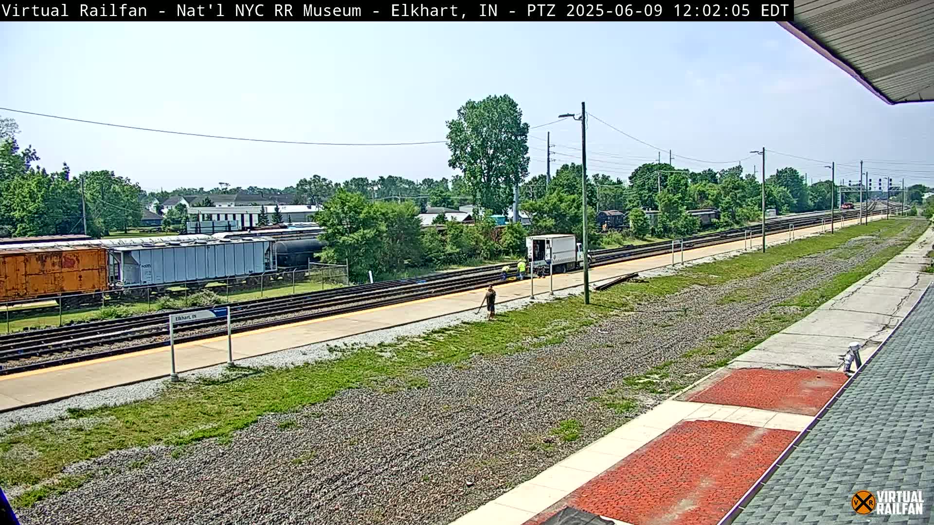A sunny day at a train station shows several railcars, workers, and a white truck next to a railway with grassy areas and buildings in the background.