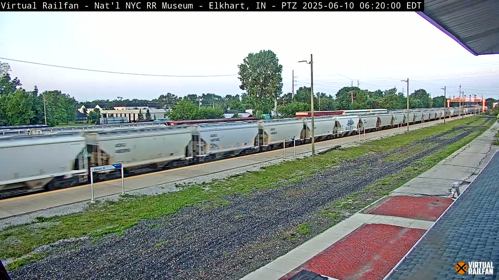 A long freight train of light gray boxcars moves past a train platform on a partly sunny day.