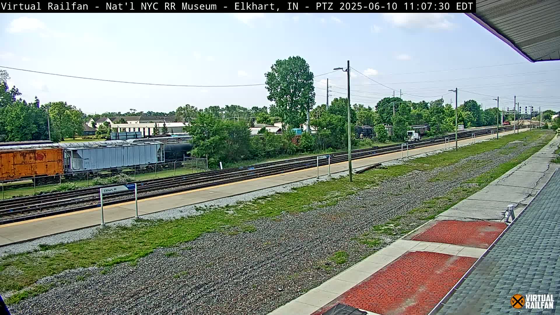 A mostly sunny view of a train station with several train cars and tracks, surrounded by green vegetation.