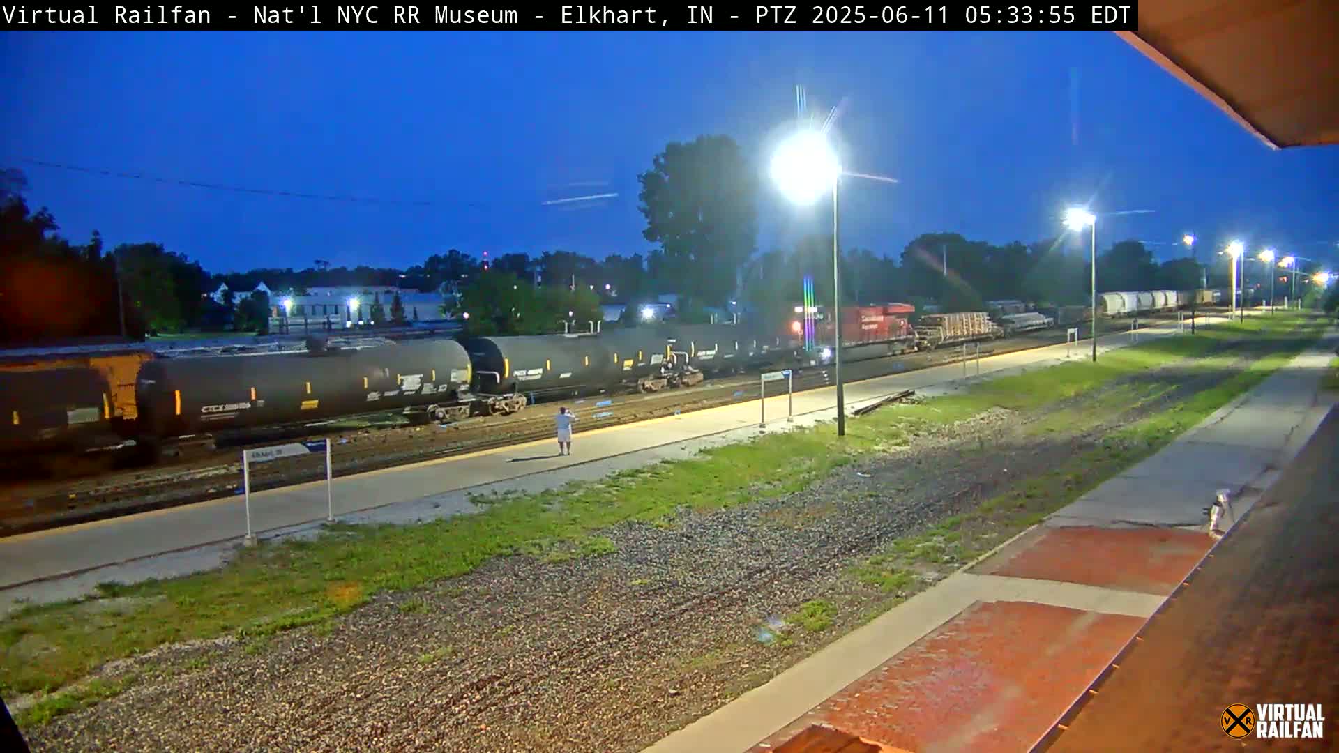 A long freight train is parked at a station at night under a twilight sky, with a single person standing nearby.