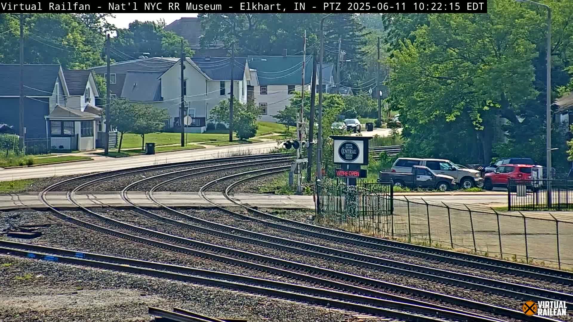 A sunny day shows several sets of train tracks curving through a residential area near a street intersection, with several cars parked nearby.