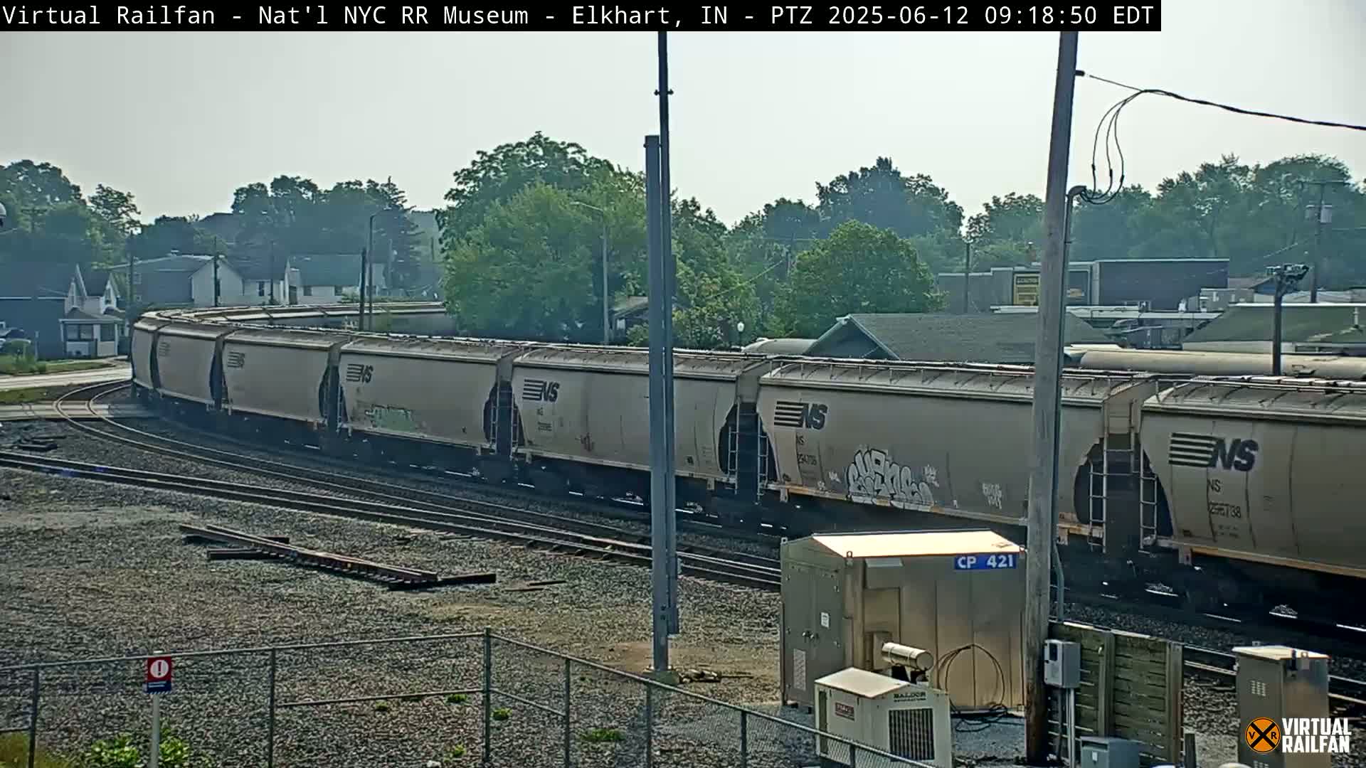 A long freight train curves around a bend in the tracks on an overcast day, passing by some industrial buildings and residential houses.