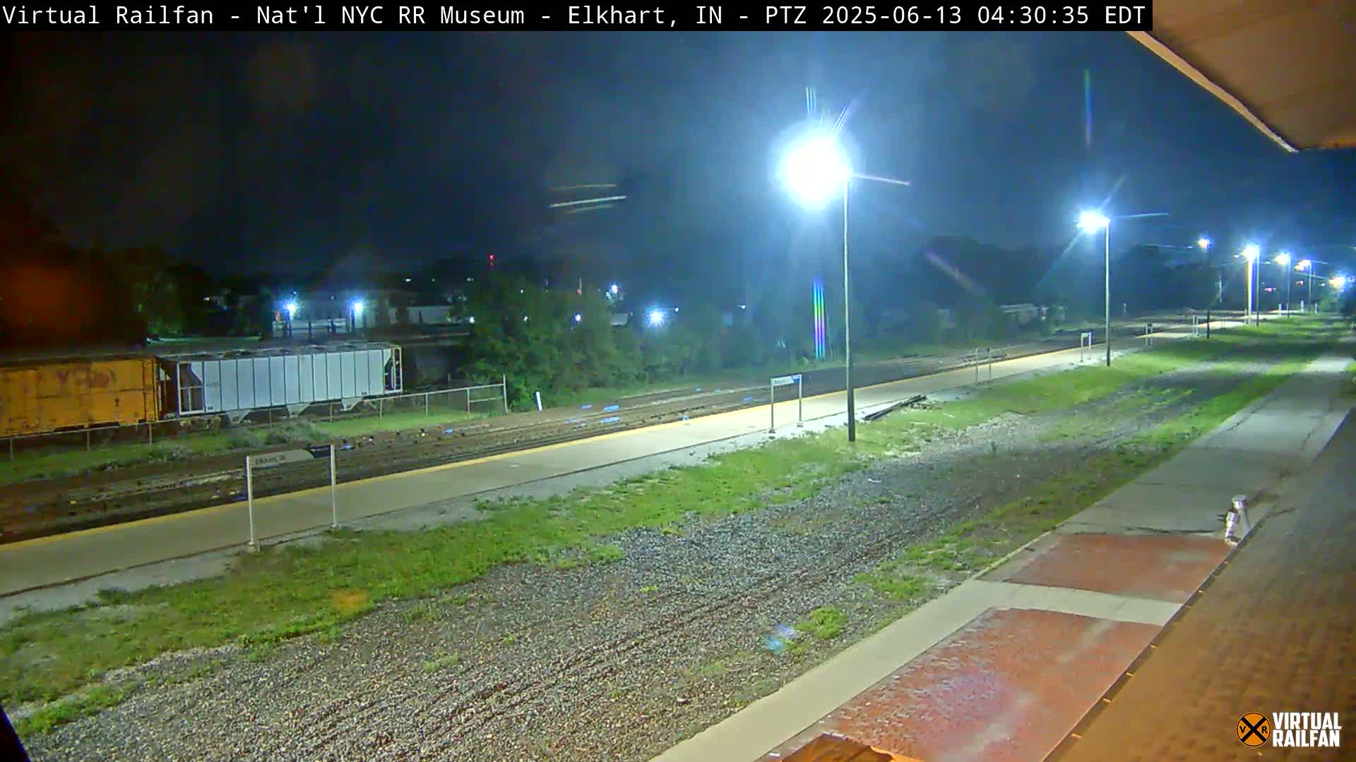A nighttime view of a train station platform with railway tracks, a few rail cars, and several lights illuminating the scene.