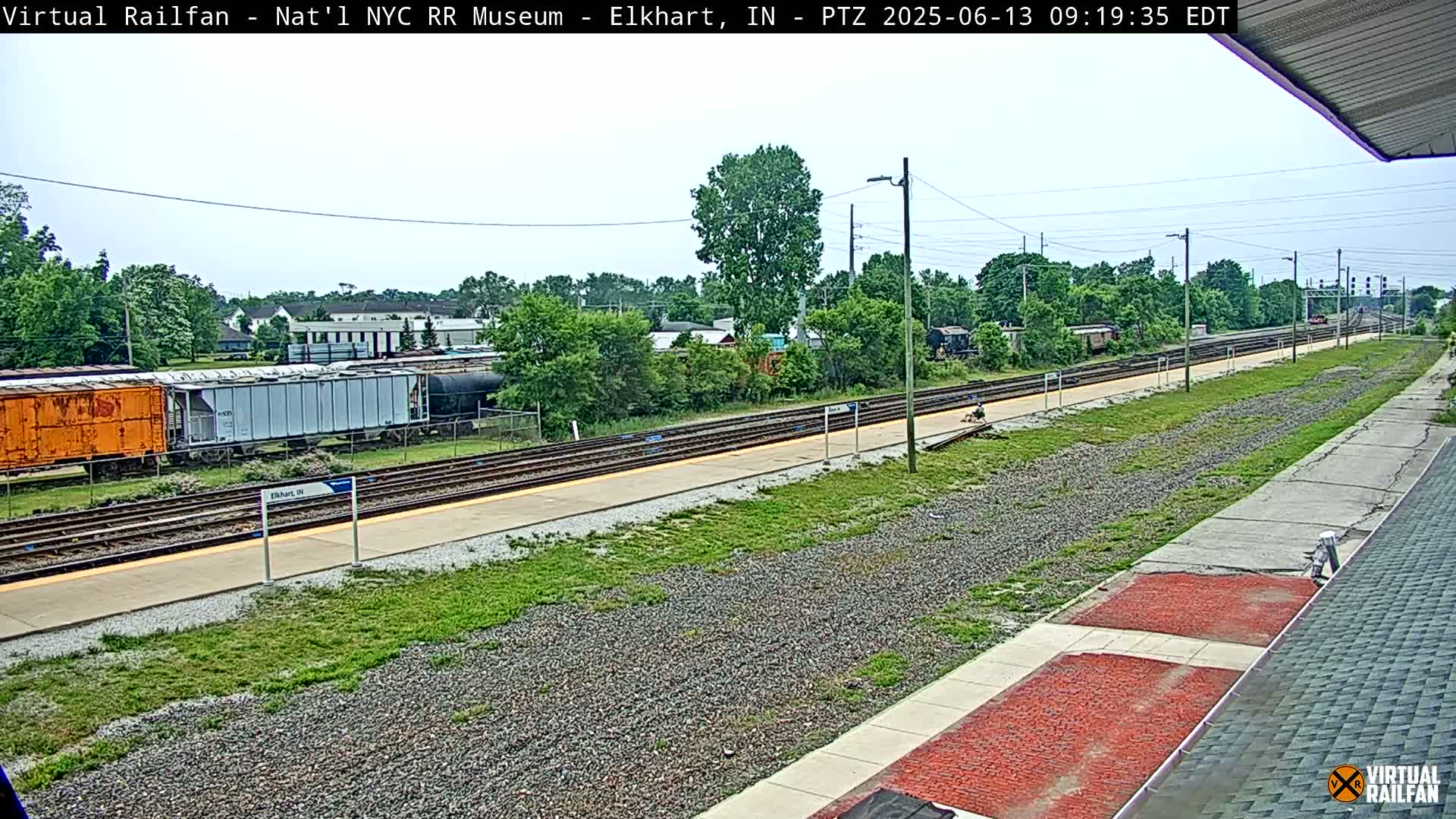 A train station platform with several railway tracks, freight cars, and surrounding greenery under a partly cloudy sky.