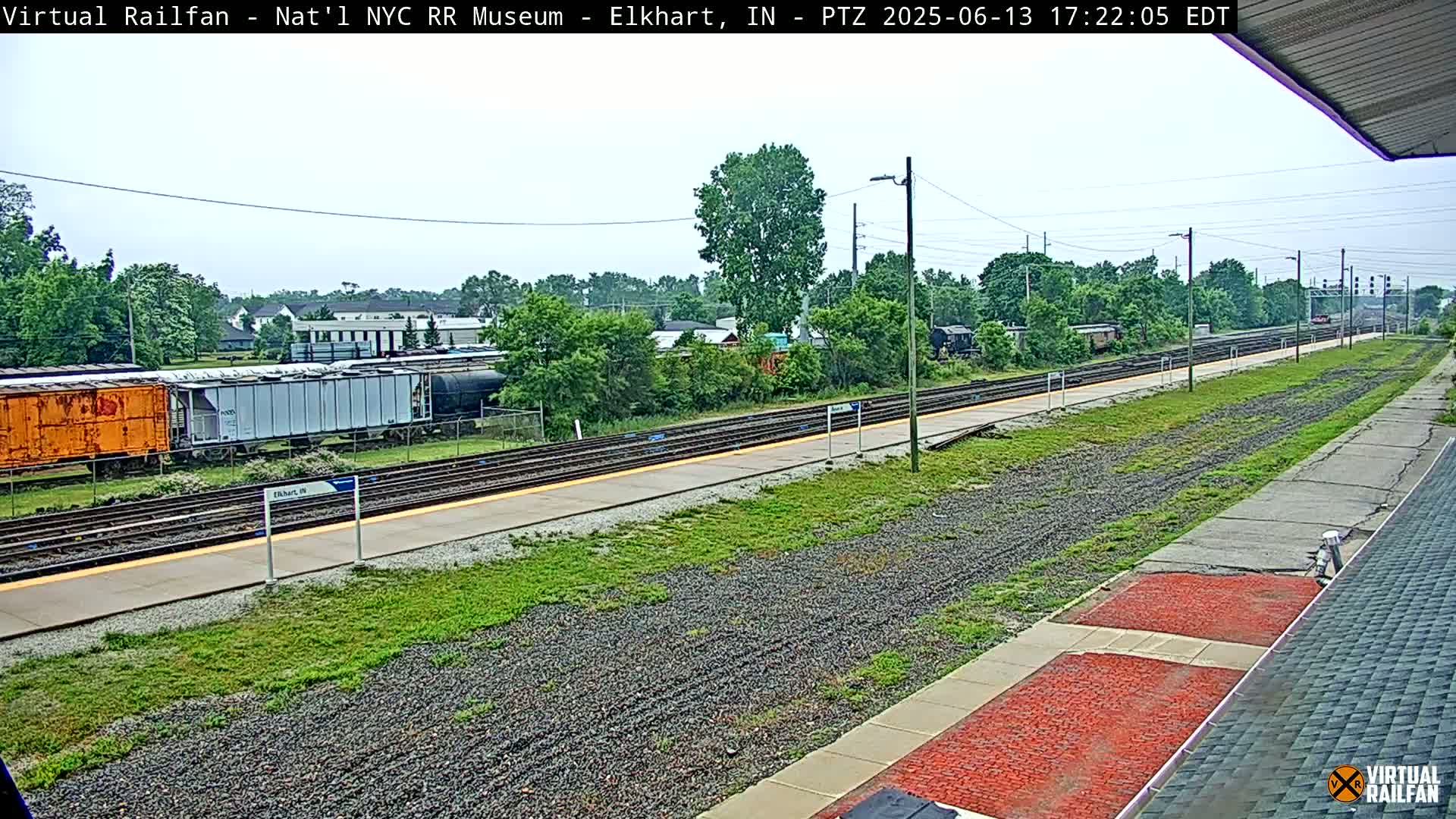 A train station platform with multiple train tracks, freight cars, and lush green vegetation under a partly cloudy sky.
