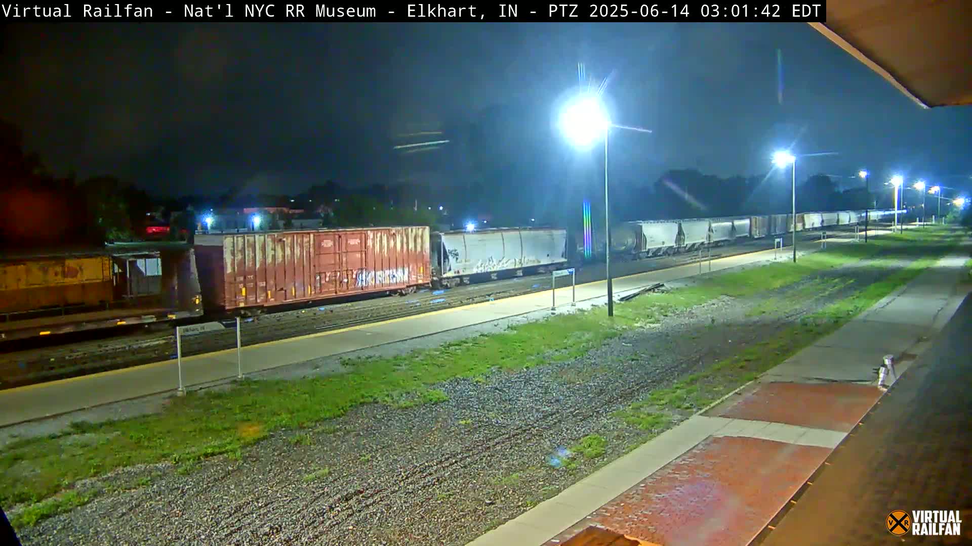 A long freight train sits at a station at night under bright lights.