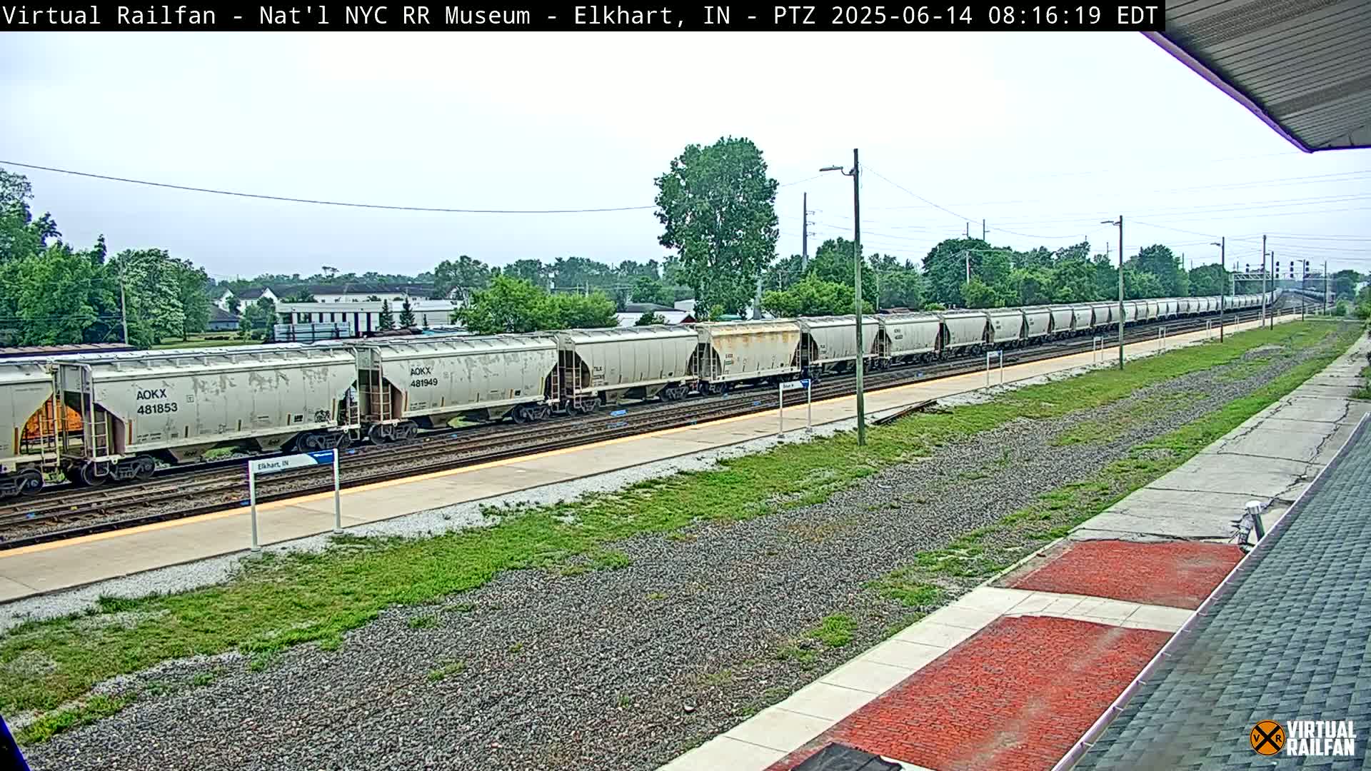 A long freight train of light gray hopper cars sits on a railway track on an overcast day, next to a grassy area and platform.